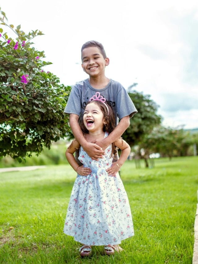 Smiling siblings in a park with vibrant greenery and colorful flowers, enjoying a sunny day outdoors.