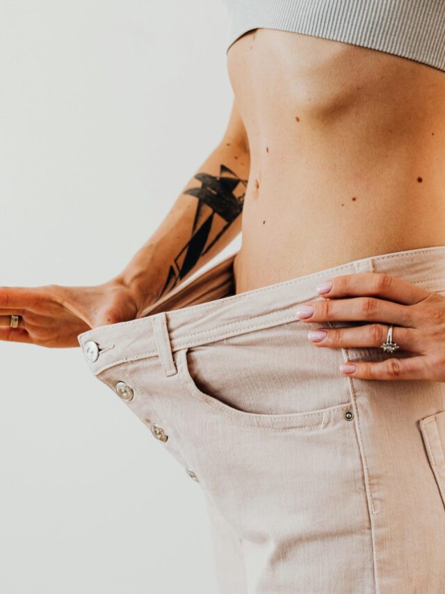 Close-up of a woman in oversized pants showcasing weight loss progress. Studio shot with neutral background.