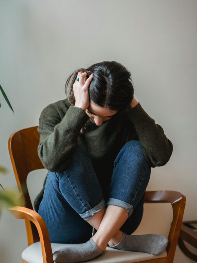 A young woman sitting on a chair indoors, holding her head in distress.