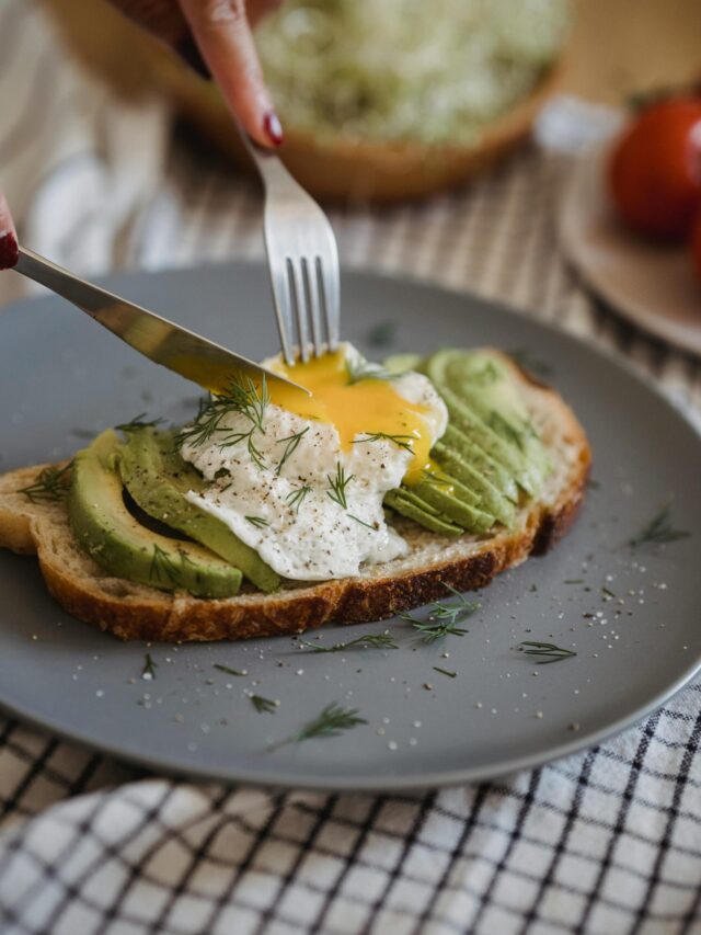 A close-up of avocado toast topped with egg, garnished with herbs on a grey plate.