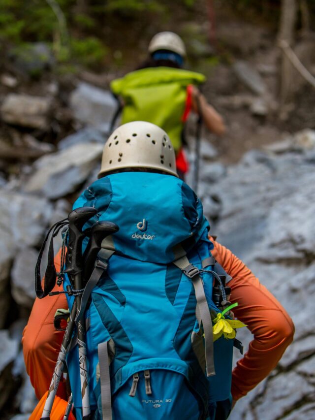 Two hikers with backpacks and helmets navigating a rocky trail in the forest.